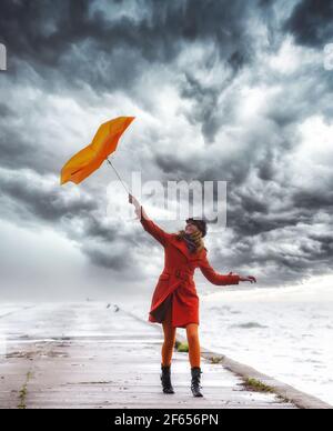 Ein Mädchen in orangefarbenem Mantel mit Regenschirm bei stürmischem Wetter auf dem Pier. Wolkiger Himmel. Stockfoto
