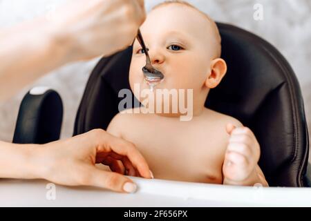 Mama füttert ein lustiges Baby aus einem Löffel, das Baby sitzt in einem Hochstuhl Stockfoto