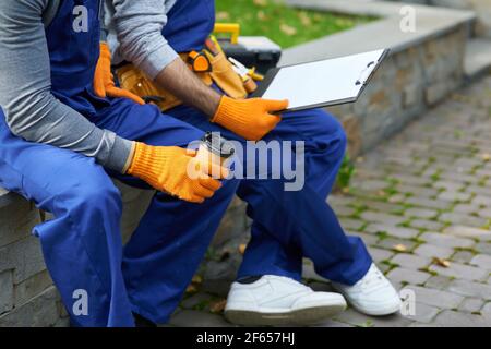 Kurzer Schuss von zwei jungen Arbeitern in Uniform sitzen im Freien Kaffee trinken während der Arbeit am Bauprojekt Stockfoto