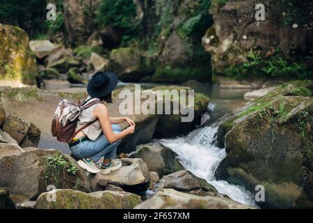 Stilvolle Frau Reisende mit Rucksack sitzen am Fluss in den Bergen. Junge Frau in lässigem Tuch und Hut auf Felsen am Fluss entspannen, Rückansicht. Reisen Stockfoto