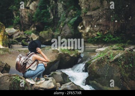 Reisen und Fernweh. Stilvolle Frau Reisende mit Rucksack sitzen am Fluss in den Bergen. Junge Frau in legeren Tuch und Hut entspannen auf Felsen bei r Stockfoto