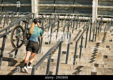 In voller Länge Schuss von professionellen Radfahrerin in Radbekleidung und Schutzausrüstung mit Fahrrad auf der Schulter zu Fuß Schritte, Training Stockfoto
