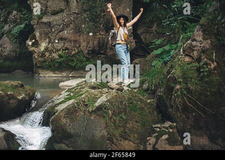 Glückliche Frau Reisende mit Rucksack stehen auf Felsen des Flusses in den Bergen. Junge Frau in legerem Tuch und Hut, die Hände am Fluss hochhält, Erfolg. T Stockfoto