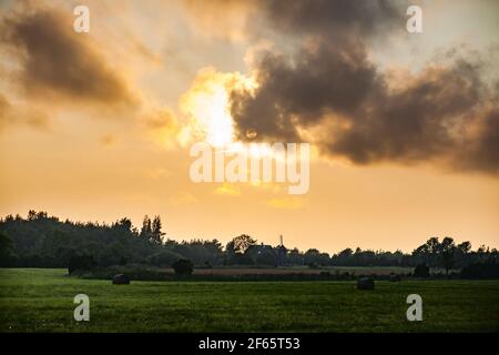 Silhouette von Landhäusern und Windmühle bei Sonnenuntergang mit Wolken und Sonne. Ländliche Landschaften Estlands. Insel Saaremaa. Stockfoto