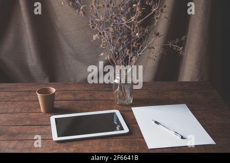 Moderner Büroarbeitsplatz mit Tablet-pc auf dem Tisch. Auf dem Tisch steht ein Tablet-Computer mit einem Blatt weißem Papier, getrockneten Blumen in einem Glas und einem DIS Stockfoto