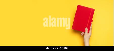 Roter Papiernotizblock in weiblicher Hand auf gelbem Hintergrund. Draufsicht femininer Arbeitsplatz. Draufsicht, flach liegend. Stockfoto