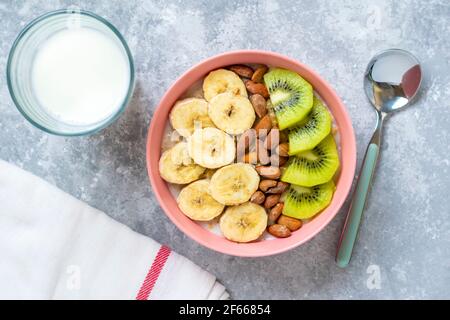 Haferflocken mit Banane, Mandeln Nüsse und Kiwi Scheiben in rosa Schüssel, Glas Milch, Serviette auf grauem Beton Hintergrund Draufsicht gesundes Frühstück oder Mittagessen Stockfoto