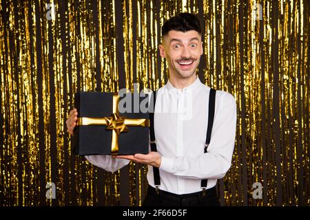 Foto von lustig aufgeregt jungen Mann tragen weißes Hemd halten Schwarze Geschenk-Box lächelnd isoliert goldenen Glitzer Hintergrund Stockfoto