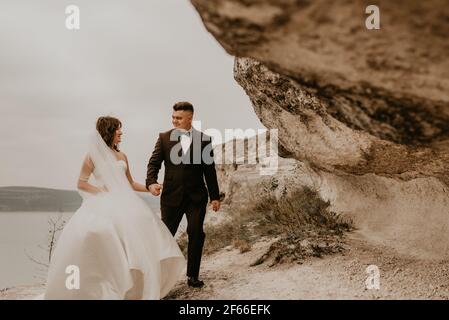 Liebevolle Paar Hochzeit frisch vermählt im Freien. Braut in weißem Kleid langen Schleier und Bräutigam in Anzug Spaziergang im Sommer fallen auf Berg über dem Fluss. sonnenaufgang. Mann ein Stockfoto