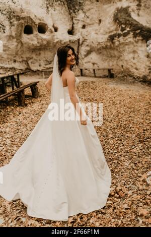 Junge Frau Braut in weißen Brautkleid mit langen Schleier laufen weg durch Herbstwald auf gefallenen orangefarbenen Blättern in den Berg. Brunette mit kurzen Haaren b Stockfoto