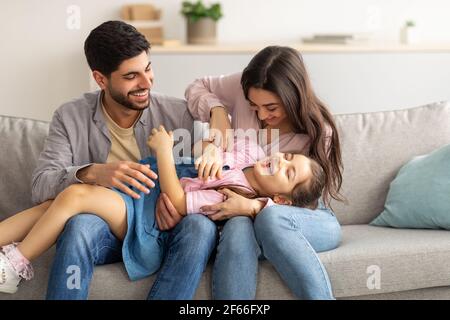 Freizeit mit der Familie. Liebevolle Eltern kitzeln ihre Tochter. Überglücklich junger Vater, Mutter und Mädchen, die Spaß zu Hause haben Stockfoto