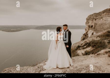 Liebevolle Paar Hochzeit frisch vermählt im Freien. Braut in weißem Kleid langen Schleier und Bräutigam in Anzug Spaziergang im Sommer fallen auf Berg über dem Fluss. sonnenaufgang. Mann ein Stockfoto