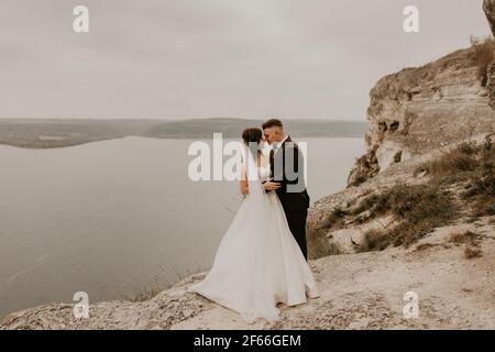 Liebevolle Paar Hochzeit frisch vermählt im Freien. Braut in weißem Kleid langen Schleier und Bräutigam in Anzug Spaziergang im Sommer fallen auf Berg über dem Fluss. sonnenaufgang. Mann ein Stockfoto