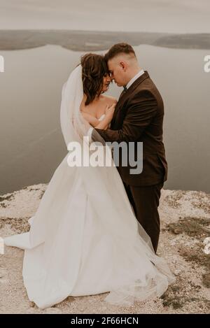 Liebevolle Paar Hochzeit frisch vermählt im Freien. Braut in weißem Kleid langen Schleier und Bräutigam in Anzug Spaziergang im Sommer fallen auf Berg über dem Fluss. sonnenaufgang. Mann ein Stockfoto