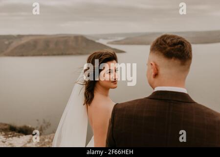 Liebevolle Paar Hochzeit frisch vermählt im Freien. Braut in weißem Kleid langen Schleier und Bräutigam in Anzug Spaziergang im Sommer fallen auf Berg über dem Fluss. sonnenaufgang. Mann ein Stockfoto