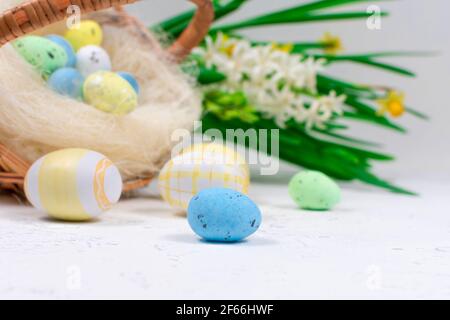 Bunte Ostereier in Pastellfarben auf einem weißen Tisch vor dem Hintergrund eines Korbes mit Ostereiern und Blumen aus Narzissen. Selektiv Stockfoto