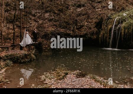 Braut in Kleid und langen Schleier Bräutigam im Anzug stehen auf hölzernen Pier über Wasser. Kleiner Teich See Fluss. Sommer-Herbst Sonnenuntergang. Wasserfall Burbun im Dorf Stockfoto