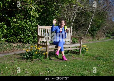 Eine Frau mittleren Alters sitzt auf einer Parkbank nach dem Training. Tasse Kaffee in der Hand und winkt einem Freund zu. Stockfoto