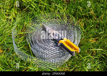 Auf dem Gras liegt eine Rolle verstärkter Maschen für Konstruktion und Drahtschneider. Gartenbau zum Schutz der Wurzeln des Sämlings vor Maulwürfen. Stockfoto