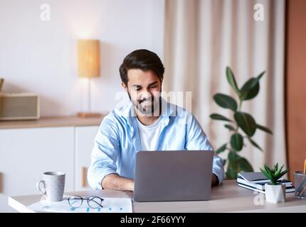 Fröhlicher Mann Aus Dem Osten, Der Im Home Office Am Schreibtisch Sitzt Und Am Laptop Arbeitet Stockfoto