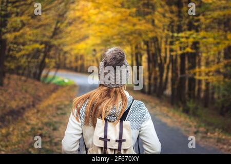 Wanderfrau trägt Strickpullover, Hut und Rucksack ist auf der Straße im Herbstwald zu Fuß. Junge Touristen suchen Wald. Reisekonzept Stockfoto