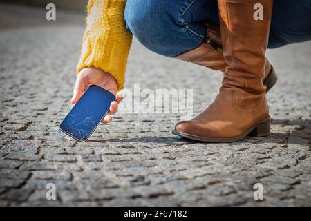 Frau, die ein kaputtes Smartphone von der Straße abholt. Gesprungener Touchscreen auf dem Mobiltelefon. Beschädigtes Mobiltelefon Stockfoto