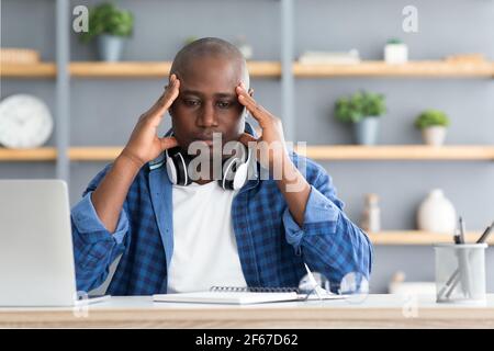 Migräneanfall. Afroamerikanischer Mann, der am Arbeitsplatz unter Kopfschmerzen leidet, während er am Laptop arbeitet Stockfoto