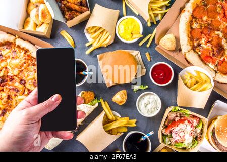 Lieferung Fastfood Bestellung von Lebensmitteln online Konzept. Große Auswahl an verschiedenen Speisen zum Mitnehmen, mans Hände mit Smartphone in pic Flatlay Rop Ansicht Stockfoto