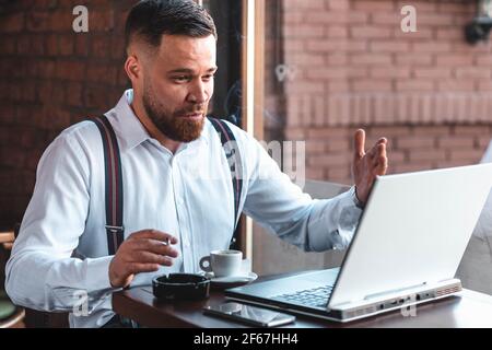 Junger Hipster-Geschäftsmann mit einem Videoanruf in einem Café, während er raucht und Kaffee trinkt Stockfoto