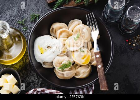 Fleischknödel, Pelmeni serviert mit Butter, Dill und Sauerrahm in schwarzer Schüssel, Draufsicht. Russische Küche Stockfoto