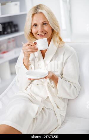 Nahaufnahme ältere kaukasische Frau lächelt in weißem Bademantel, hält Tee im Kosmetiksalon Stockfoto