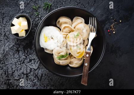 Pelmeni, Fleisch gefüllte gekochte Knödel mit Butter, Dill und Sauerrahm auf schwarzem Hintergrund, Draufsicht Stockfoto