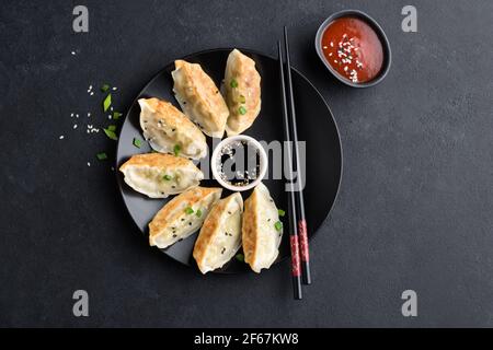 Asiatische Küche frittierte Gyoza- oder Jiaozi-Knödel mit Sojasauce, Shriracha-Sauce und Sesamsamen auf schwarzem Beton-Hintergrund, Draufsicht Stockfoto