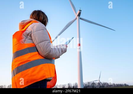 Eine Ingenieurin, die eine Tablette in der Hand hält und im Windturbinenpark auf dem Feld in der orangefarbenen vesta arbeitet Stockfoto