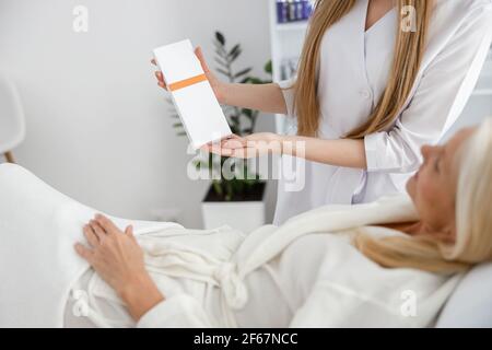 Foto einer älteren Frau im weißen Bademantel, der auf dem Tagesbett liegt Und die Überprüfung der Ergebnisse der Hautpflege Behandlung, während Kosmetologe halten Glasfassade Stockfoto