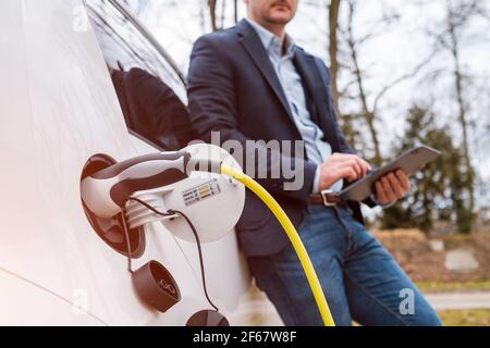 Geschäftsmann, der in der Nähe eines Elektroautos oder eines Elektroautos steht Und mit Tablet auf der Straße bei Sonnenlicht Stockfoto