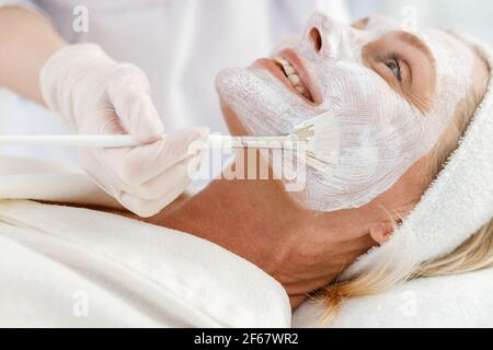 Nahaufnahme Foto der Kosmetikerin Anwendung Ton Gesichtsmaske auf Gesicht einer älteren Frau Stockfoto
