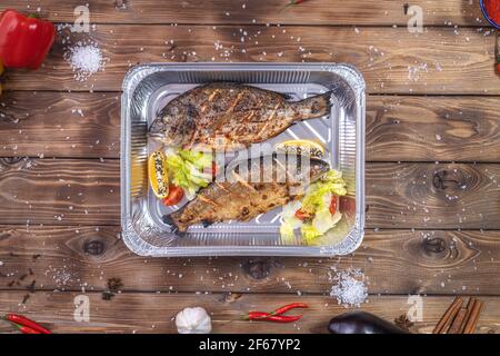 Gegrillter Fisch mit Gemüse, Paprika, grobes Salz, in einem Aluminiumbehälter auf braunem Holzgrund gelegt. Lieferung von Speisen im Restaurant. Stockfoto