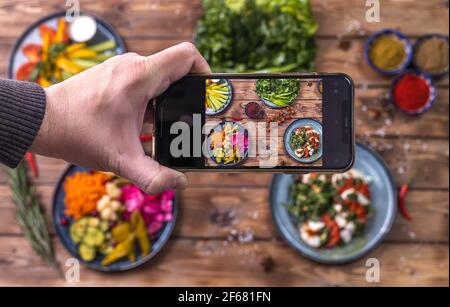 Eine Person macht Fotos von eingelegtem Gemüse auf seinem Telefon: Paprika, Karotten, Knoblauch, Kohl auf einem hölzernen Hintergrund mit Chilischoten verziert, Cherr Stockfoto