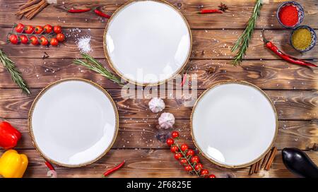 Leere Teller stehen auf einem braunen Holzgrund, der mit Kirschtomaten, Rosmarin, Chilischote und grobem Salz verziert ist. Flaches Layout. Kopieren Sie den Speicherplatz. Stockfoto