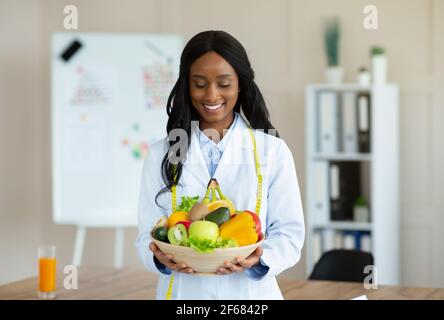 Portrait von fröhlichen schwarzen Ernährungsberater in Laborkittel halten Schüssel Von frischem Obst und Gemüse in der Gewichtsverlust Klinik Stockfoto