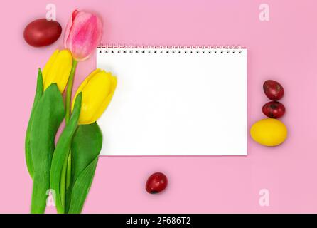 Leeres Notizbuch mit Spirale für Text, Tulpen mit farbigen Eiern auf rosa Hintergrund. Osterkarte. Speicherplatz kopieren Stockfoto