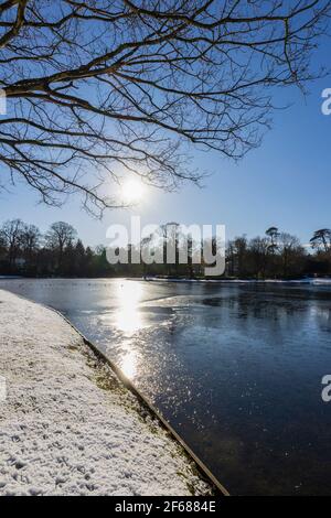 Wintersonne über dem gefrorenen See im Claremont Landscape Garden bei Esher, Surrey, Südostengland an einem verschneiten Tag Stockfoto