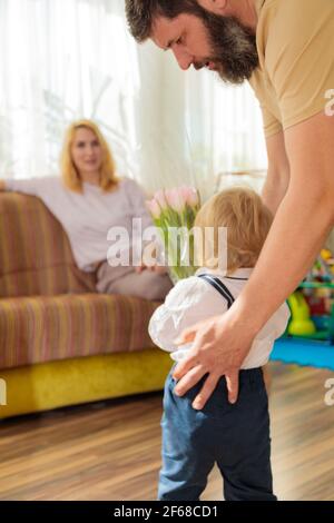 Papa mit Baby, Sohn gratuliert Mama zum Urlaub. Das Kind bringt seiner Mutter einen Blumenstrauß und ein Geschenk. Stockfoto