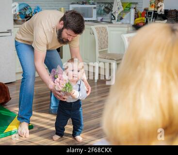 Papa mit Baby, Sohn gratuliert Mama zum Urlaub. Das Kind bringt seiner Mutter einen Blumenstrauß und ein Geschenk. Stockfoto