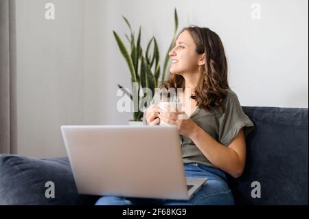 Freiberuflerin beim Träumen mit einer Tasse Kaffee am Morgen, lächelnde Frau sitzt auf der Couch mit einem Laptop, schaut weg, wartet auf Inspiration oder neue Ideen Stockfoto