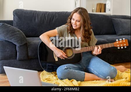 Hobby und Freizeitgestaltung Konzept. Junge attraktive Frau spielt Gitarre mit Video-Tutorial auf dem Laptop, sitzt Frau auf dem Boden vor dem Computer hält akustische Gitarre und lernen, wie man spielt Stockfoto