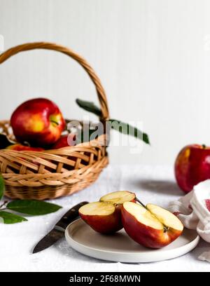 Frisch gepflückte, leckere, reife und süße rote Äpfel liegen in der Küche, einer wird geschnitten, um als gesunder Snack genossen zu werden, einige noch im Korb. Stockfoto