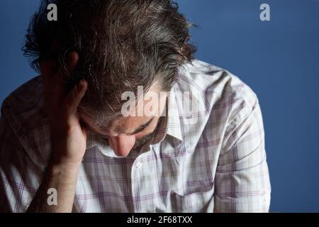 Junger Kaukasier mit gestreiftem Hemd sitzt, während er nach unten schaut und seinen Kopf mit der rechten Hand gegen hellblauen Hintergrund hält. Stockfoto