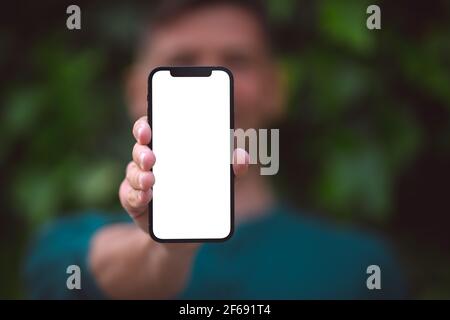 Mann hält modernes Smartphone mit Mockup-Bildschirm oder leeren Bildschirm. Junger Mann zeigt sein Telefon mit leerem Raum im Freien auf grünem Hintergrund Stockfoto